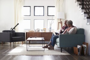 An older black couple sitting on a light blue couch in a light and airy room reading a brochure together. There is a table in front of them and a modern feel to the furniture
